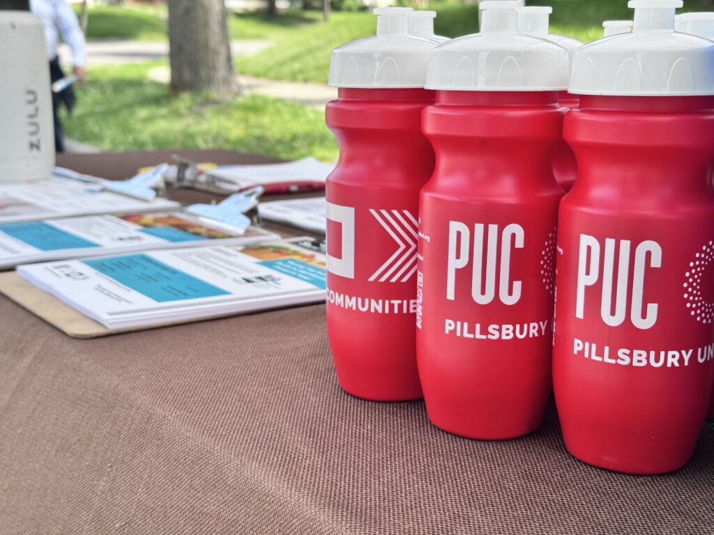Red water bottles with the Pillsbury United Communities Logo rest on a table. Next to them is a collection of clipboards with community resource flyers on them.